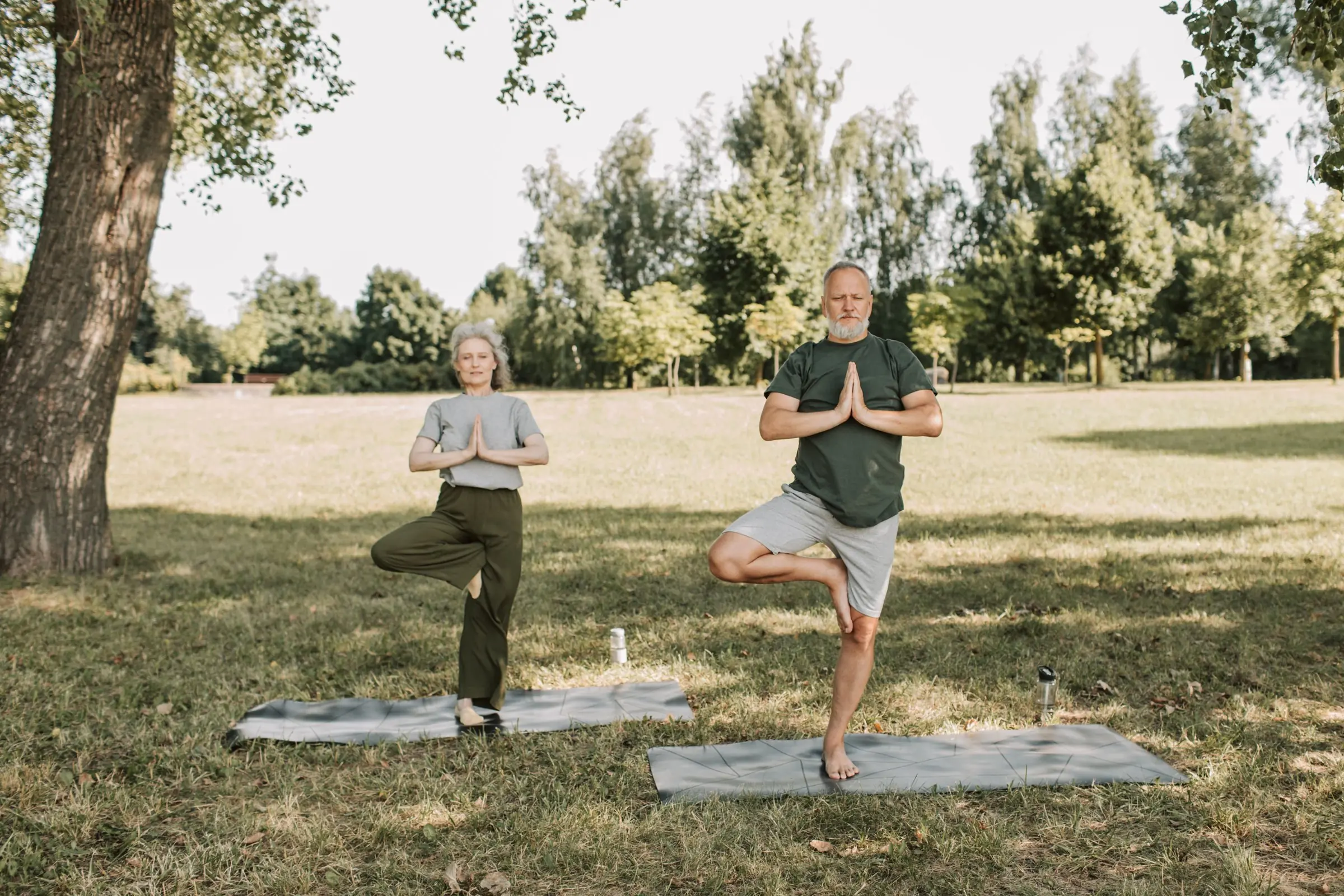 An older man and woman standing on one leg in a balance pose on a park lawn