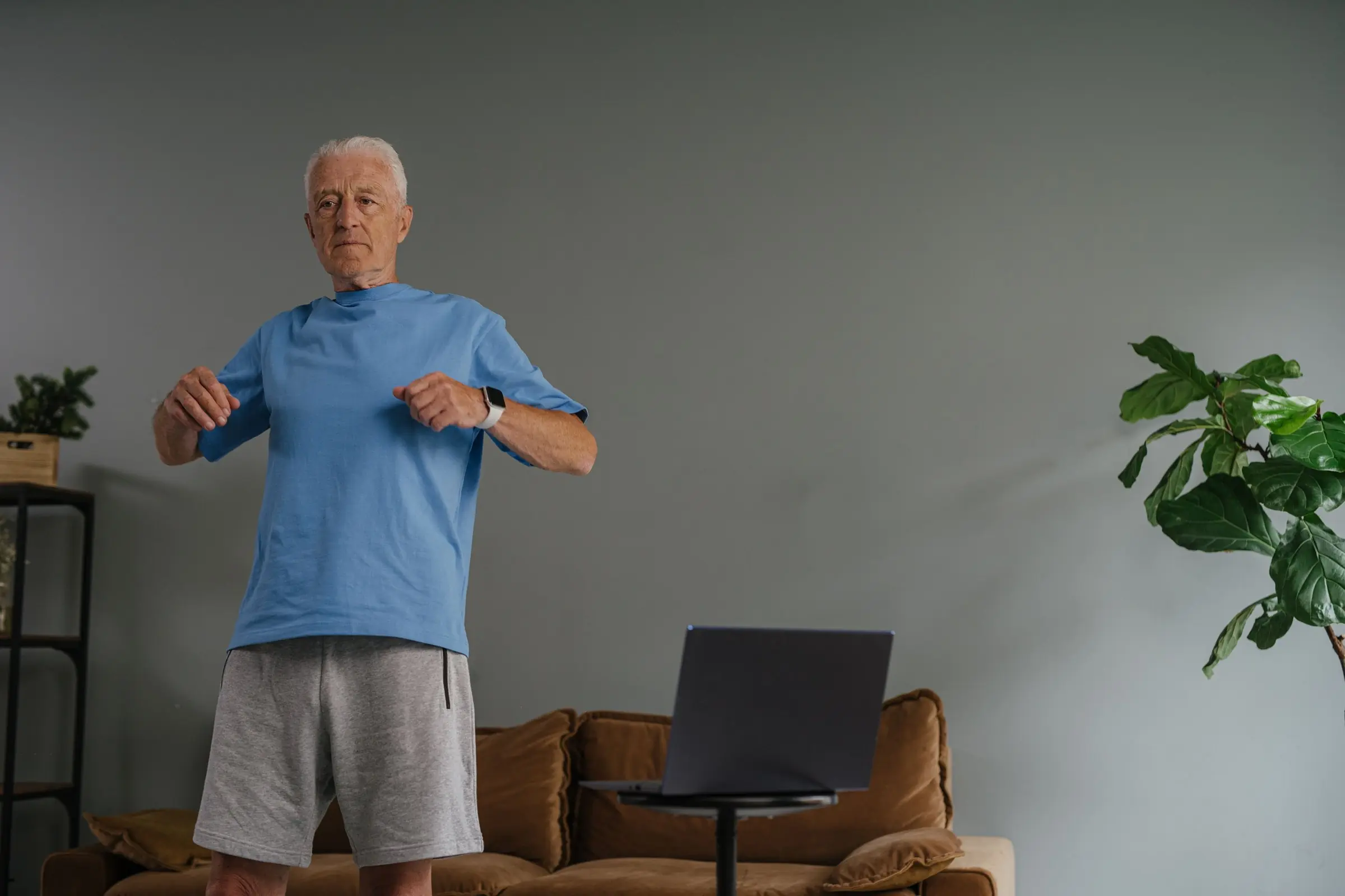 An older man doing a standing exercise at home in front of a laptop