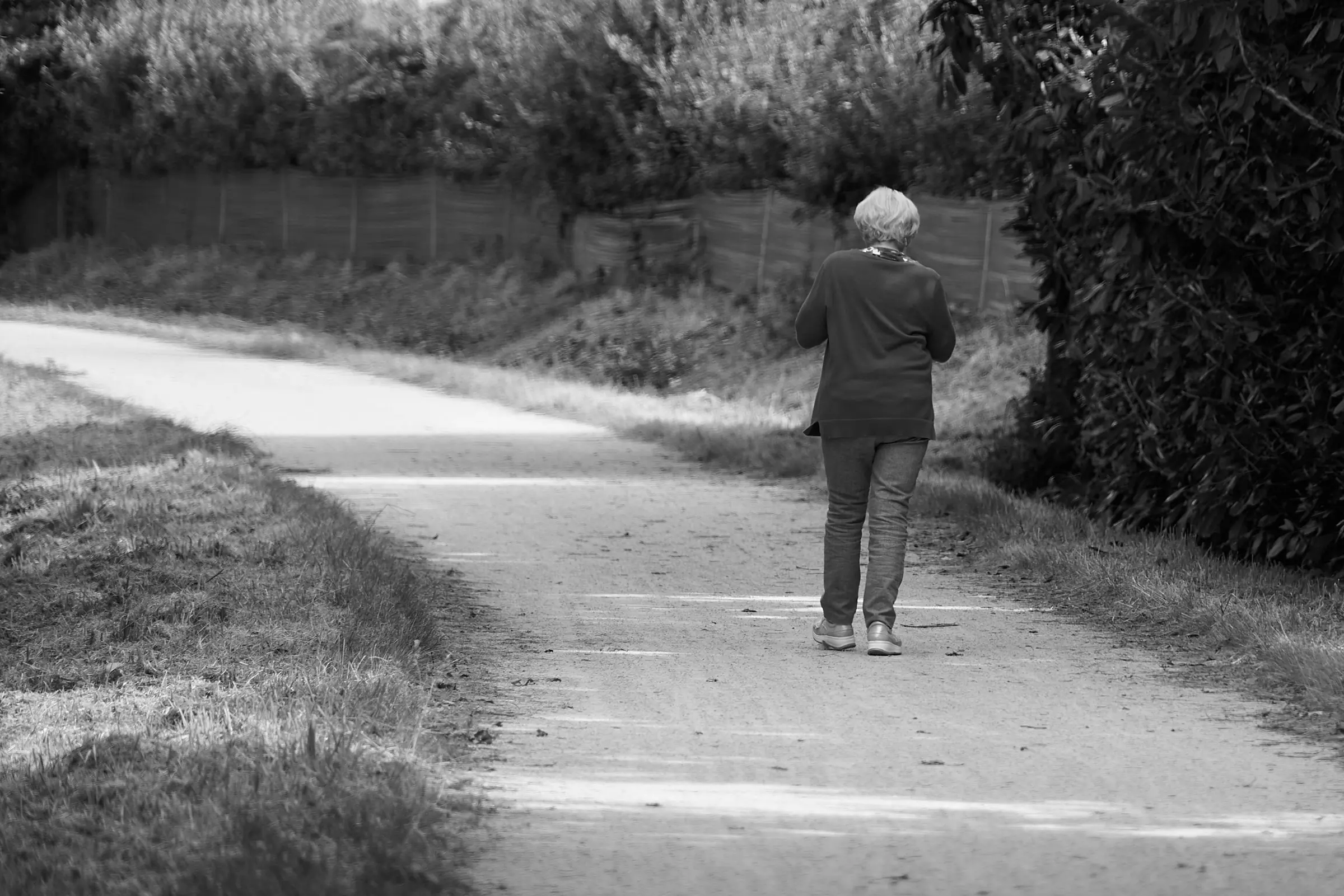 A black and white photograph of an older woman walking alone down a quiet country path